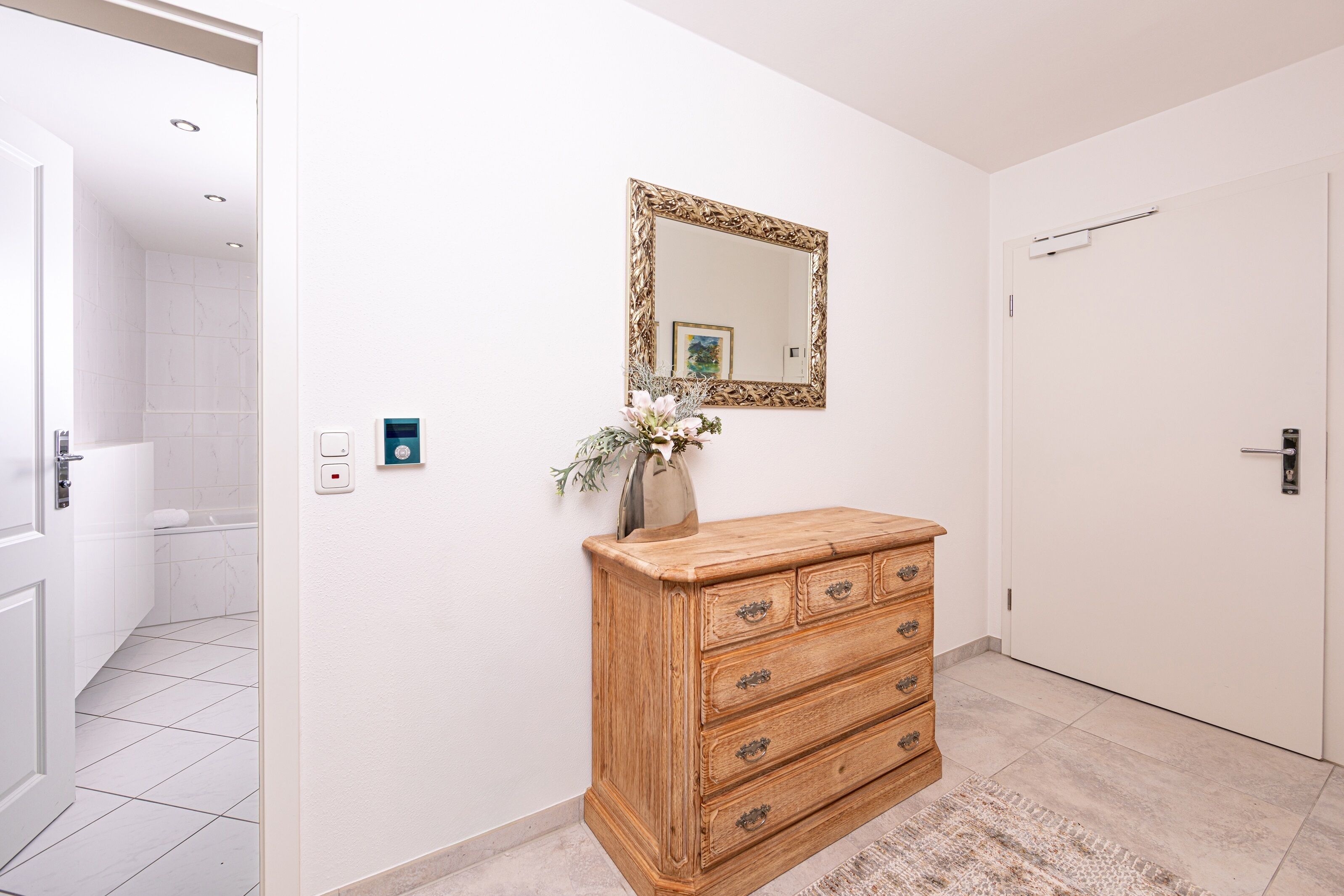 Un baño con azulejos blancos, un aparador de madera con cajones, un espejo con marco decorativo y una puerta con picaporte.