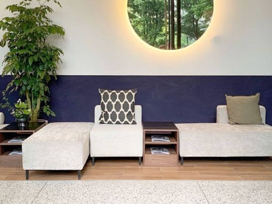 A reception area with a round wall-mounted light, a potted plant, two sofas with patterned cushions, and a doormat with the word 'base'.