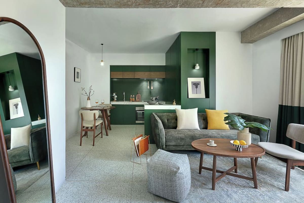 Una sala de estar moderna con sofá, reposapiés y mesa de centro, un comedor con sillas y mesa, y una cocina con gabinetes verdes e iluminación colgante.