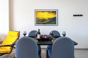 A dining area with a table set for four, a yellow chair, and a framed landscape painting on the wall. - Vox 2-BR Apt & Balcony in Larnaca (Larnaca)