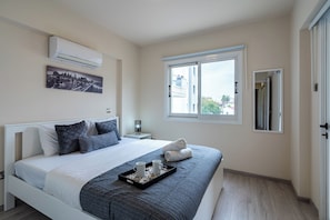A modern bedroom with a bed, a window, an air conditioner, and a cityscape painting. - Vox 2-BR Apt & Balcony in Larnaca (Larnaca)