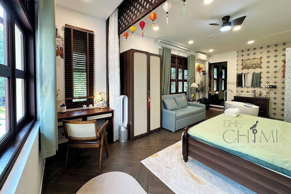 A modern bedroom with a bed, a desk, a chair, a sofa, a bathroom, and a window with blinds.