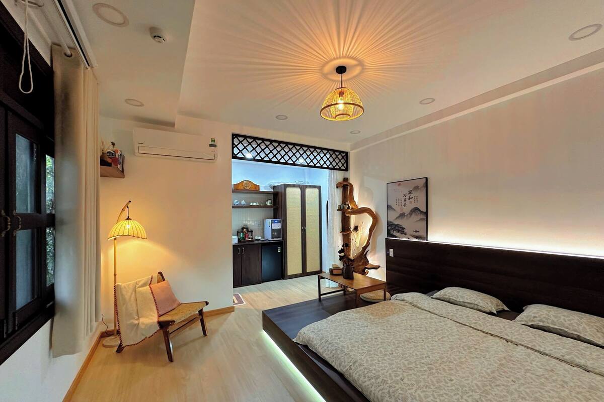 A modern bedroom with a bed, bedside table, lamp, and a view into the kitchen area.