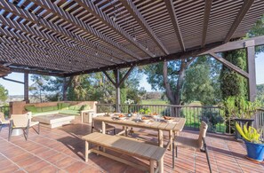 Een terras buiten met een houten eettafel, een bank en een pergola erboven.