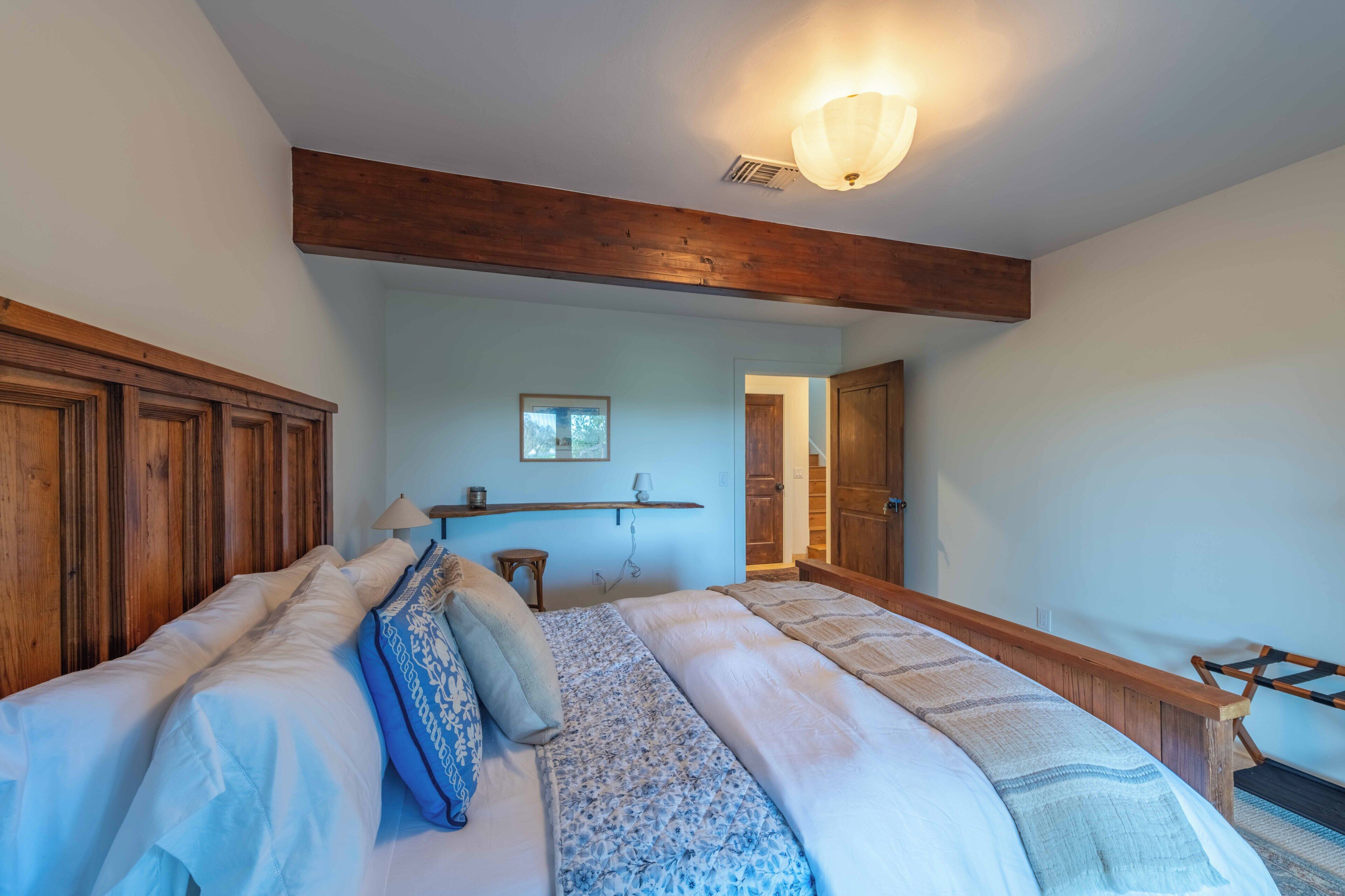 A bedroom with a wooden headboard, a bed with multiple pillows and blankets, a nightstand with a lamp, and a wooden bench by the door.