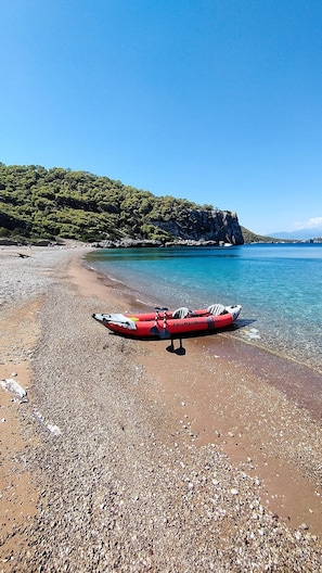 Ein rotes Luftboot an einem Kiesstrand mit klarem Wasser und einer bewaldeten Hügelkette im Hintergrund.