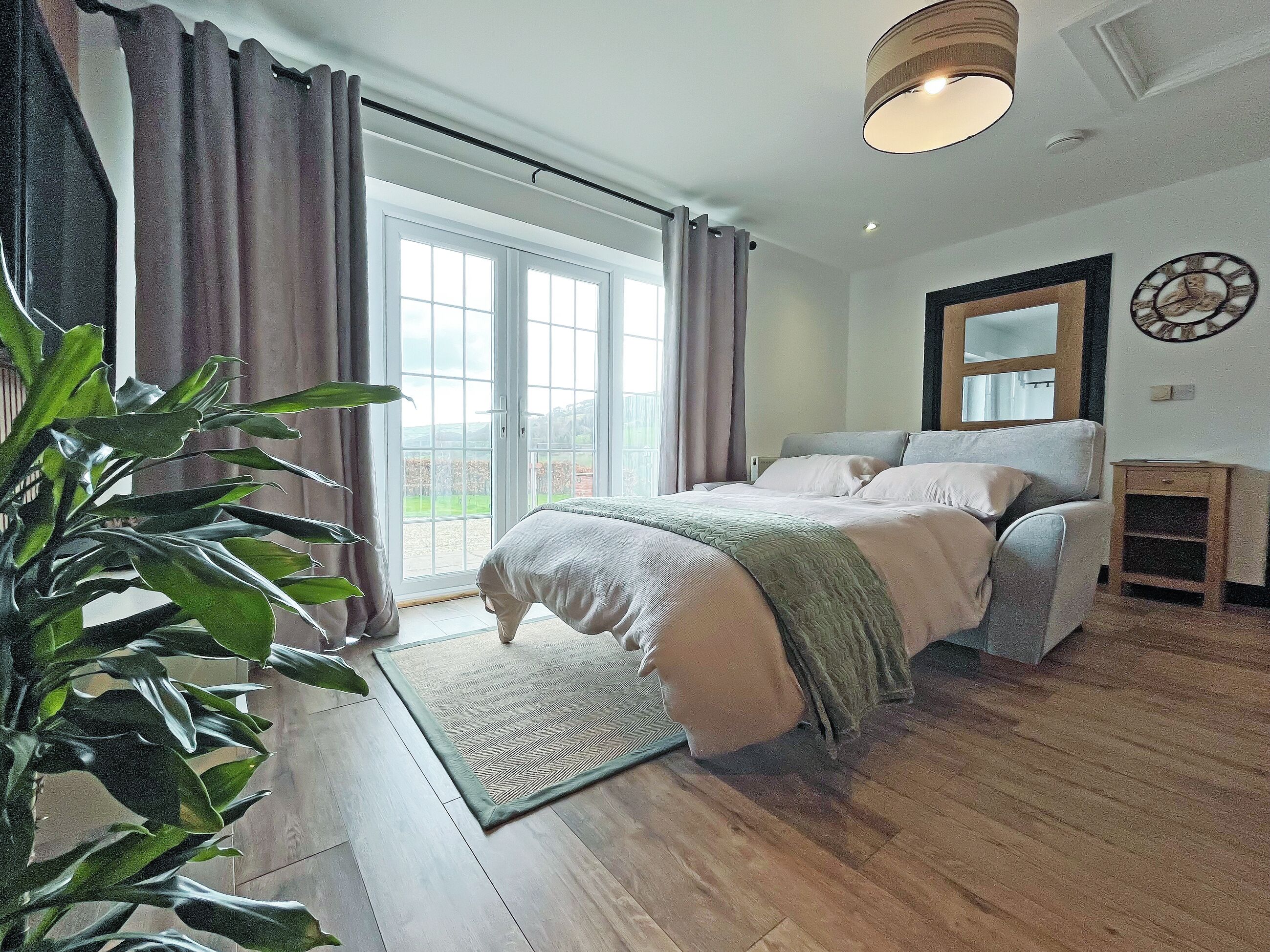 Une chambre à coucher avec un lit, une table de chevet, un miroir et une vue sur l’extérieur grâce à une grande fenêtre.