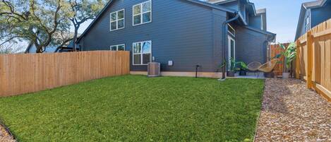 A residential backyard with a lawn, a wooden fence, a house with a dark exterior, and a small garden area with plants and a chair.