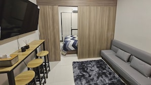 A compact living space with a TV, a sofa, a dining area with stools, and a bed with curtains.
