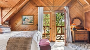 A wooden cabin bedroom with a bed, a painting, a dresser, and a view of the outdoors.