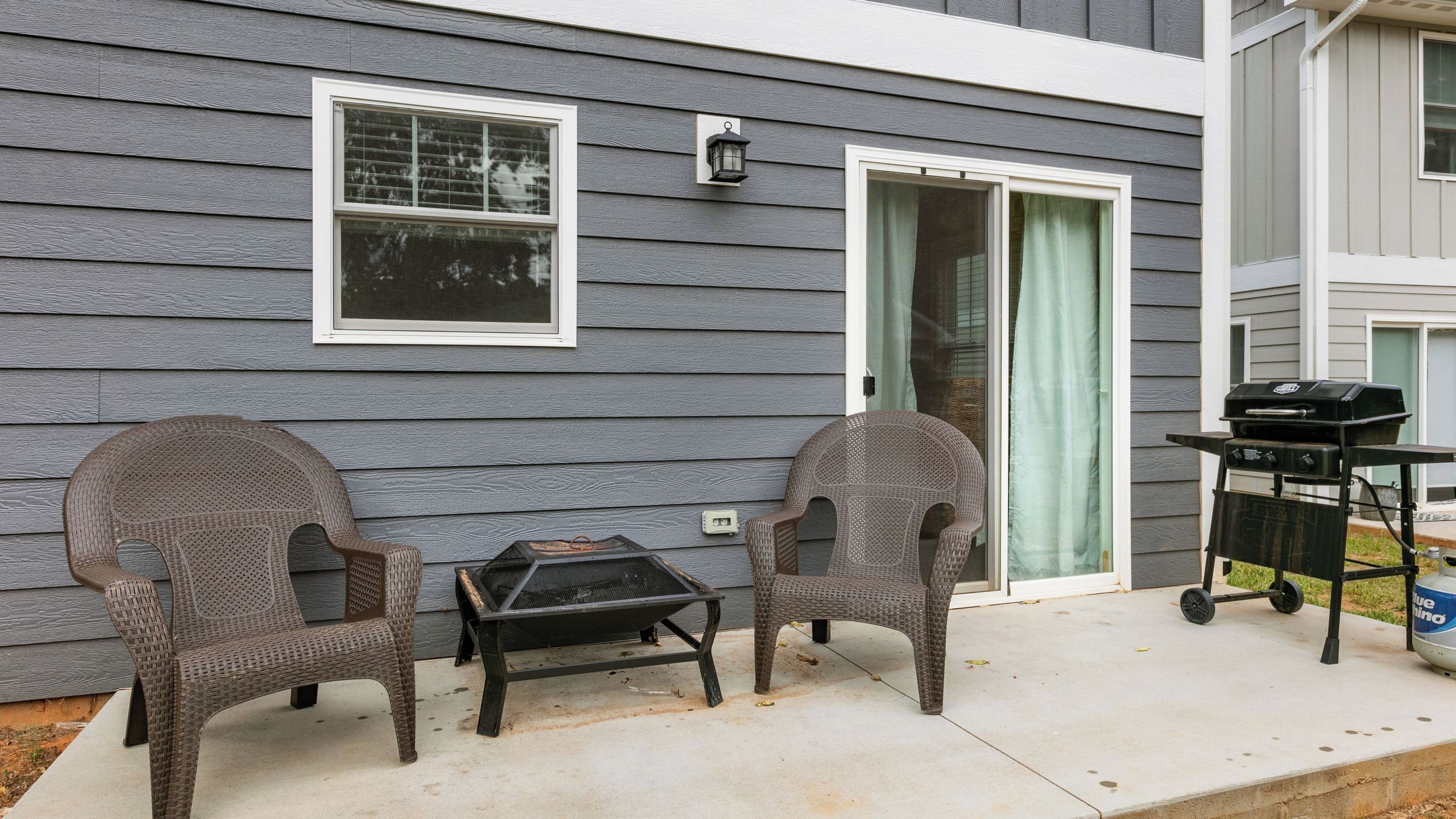 Een huis met een terras, twee rieten stoelen, een kleine tafel en een barbecue.