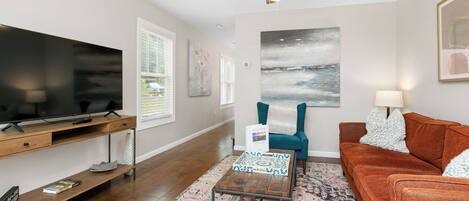A living room with a flat-screen TV, a wooden coffee table, a patterned rug, a teal armchair, and a brown sofa.