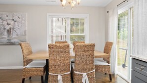 A dining area with a wooden table and wicker chairs, a painting on the wall, and a window with blinds. - Red Bud Sycamore by Avantstay Custom Group Retreat w/ Patio (Asheville)
