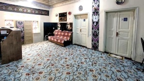 A room with a patterned carpet, a sofa, a desk, and a door with a number. - oybek hotel (Khiva)