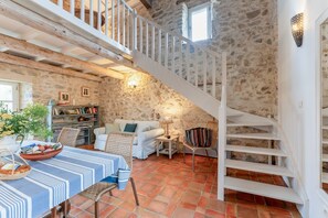 Un intérieur rustique avec un mur en pierre, un escalier blanc, un coin repas avec une table et un canapé.