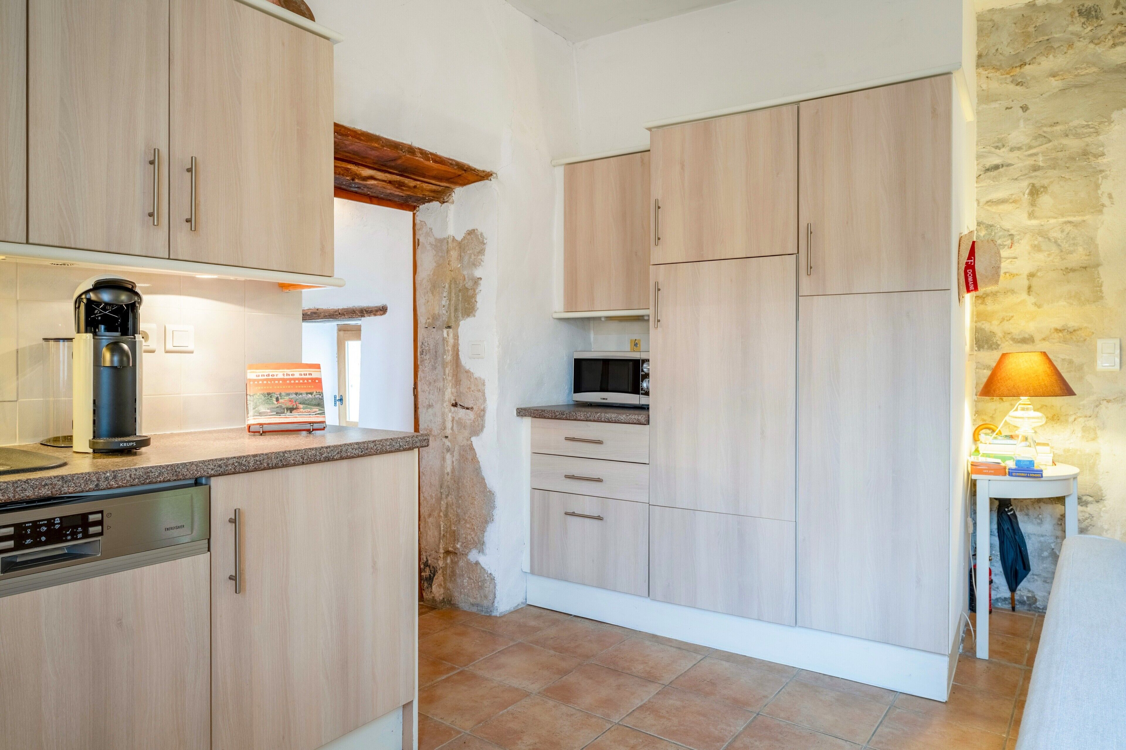 Une cuisine avec des meubles de couleur bois clair, une machine à café, un four à micro-ondes et une petite table avec une lampe.
