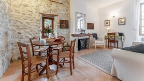 A stone-walled room with a dining table, chairs, a fireplace, a sofa, and a desk with a lamp.