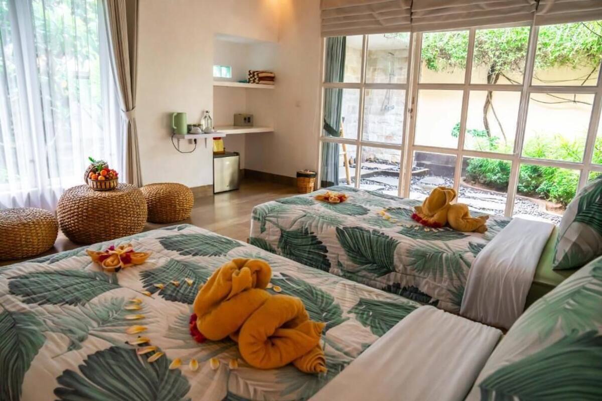 A room with two beds, a small kitchenette, and a large window with a view of greenery.