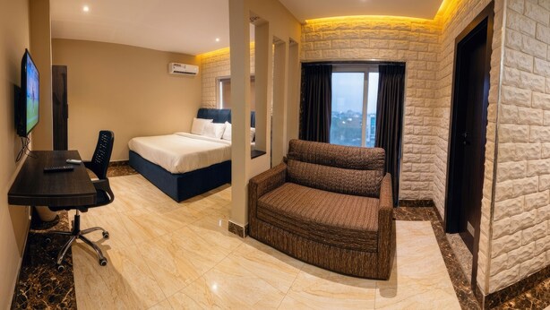 A hotel room with a bed, a sofa, a desk with a chair, a TV, and a large window with a view. - Citistay Apartments (Abuja)
