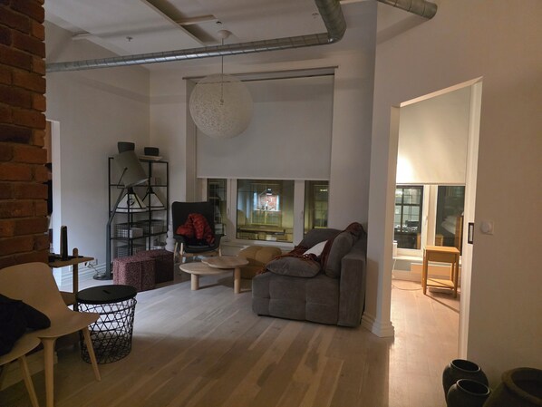 Ein modernes Wohnzimmer mit grauem Sofa, rundem Couchtisch und schwarzem Sessel. An der linken Seite eine Ziegelwand, eine große weiße Pendelleuchte und ein Blick in die Küche durch eine Glastür.