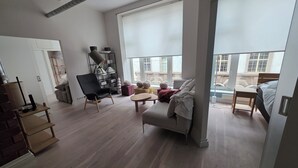 Ampla sala de estar com sofá, poltrona e uma pequena mesa. Há uma estante com objetos decorativos e uma grande janela com vista para a cidade.