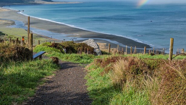 A coastal path leading to a beach with a rainbow, grassy areas, and a wooden fence.