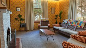 A cozy living room with a sofa, armchair, and coffee table, featuring patterned wallpaper, curtains, and potted plants.