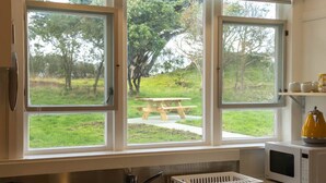 A kitchen with a sink, a microwave, and a window overlooking a grassy area with trees and a picnic table.