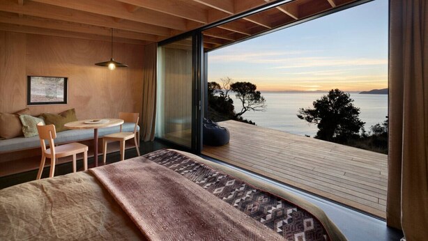 A modern room with a large window overlooking a body of water, a bed, a seating area with a table, and a view of the sunset.