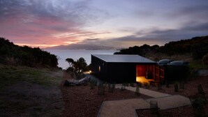 A modern cabin with a glass door, situated on a hillside overlooking a body of water.