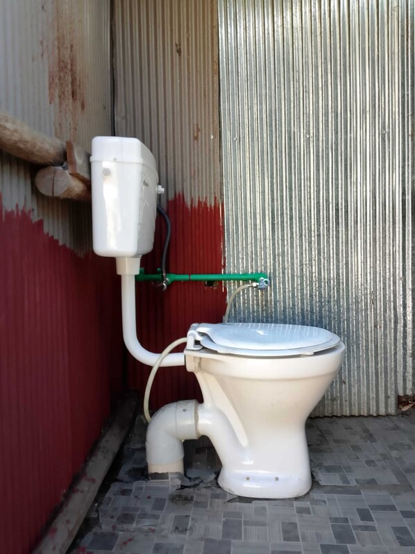 A toilet with a green pipe and a white tank, situated in a narrow space with corrugated metal walls and a tiled floor. - River View Cafe & Camping (Jibhi)