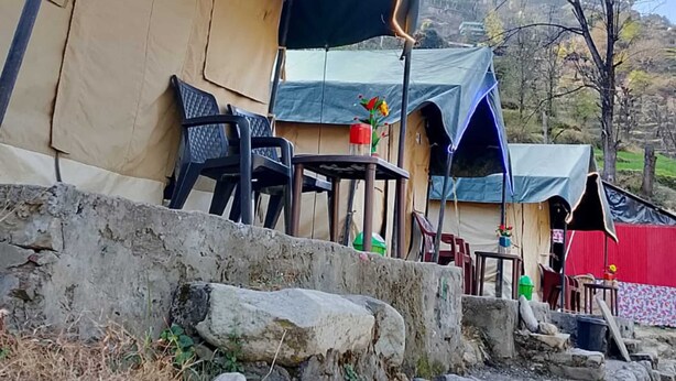 A campsite with tents, a table with chairs, and a stone wall. - River View Cafe & Camping (Jibhi)