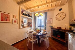 A dining area with a round table, white chairs, a chandelier, a fireplace, and framed artwork on the walls.