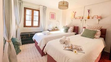 A hotel room with two beds, a window with curtains, a bedside table with a lamp, and a shelf with decorative items.