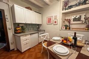 A kitchen with white cabinets, a fruit bowl, and a dining table set for two with plates, glasses, and a bottle of wine.