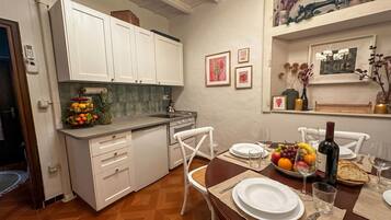 A kitchen with white cabinets, a fruit bowl, and a dining table set for two with plates, glasses, and a bottle of wine.