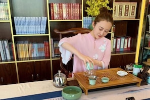 Une personne verse du thé dans une tasse sur un plateau où se trouvent une théière et d’autres tasses. On aperçoit des étagères à livres en arrière-plan.