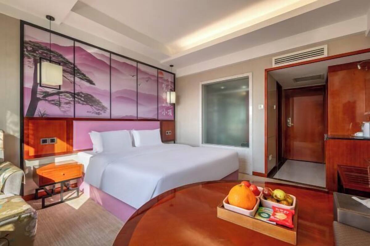 Quarto de hotel com uma cama, uma mesa de cabeceira com frutas, um quadro na parede e uma janela.