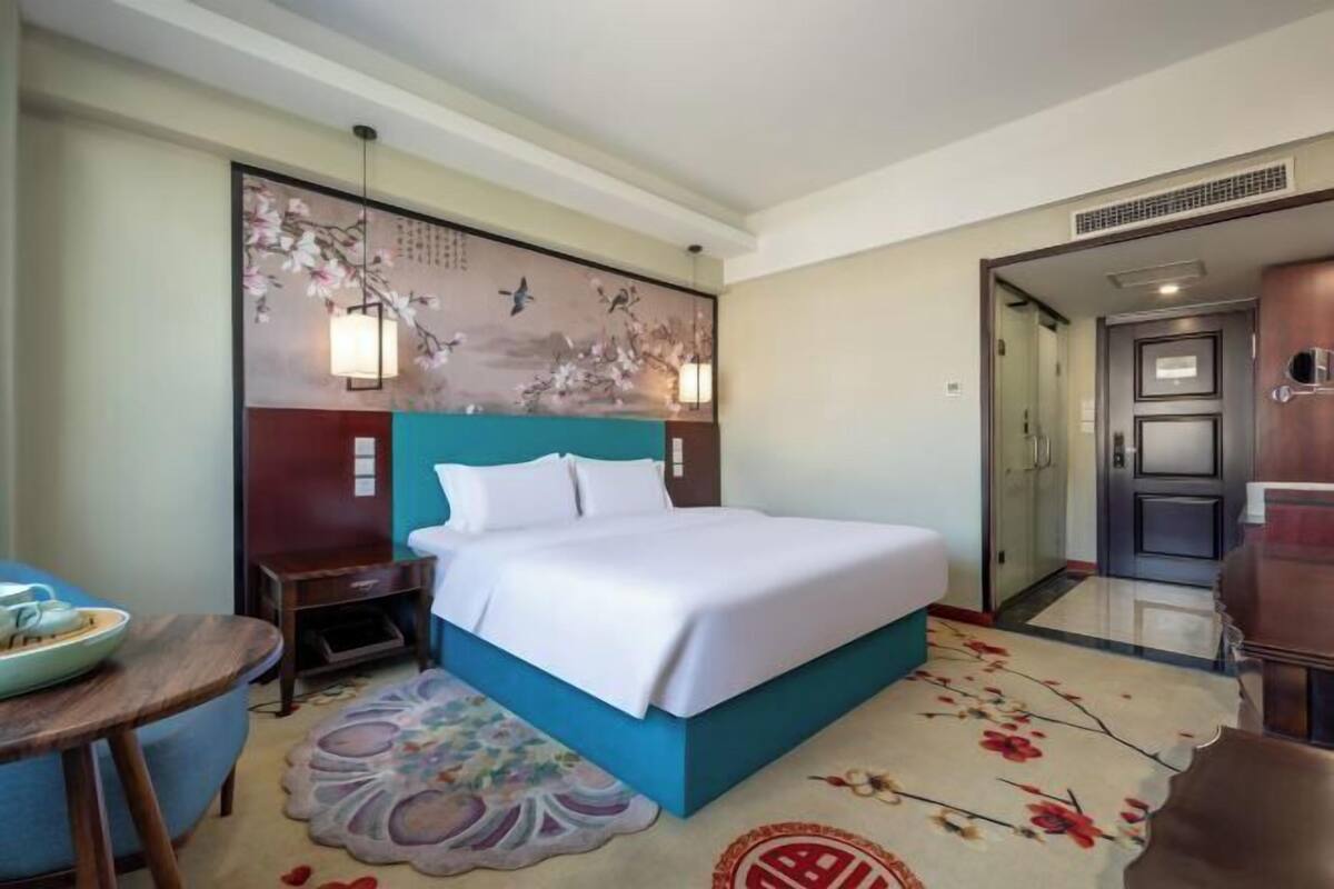 Quarto de hotel com cama, mesa de cabeceira, cadeira, parede com obra de arte e porta que dá para outro quarto.