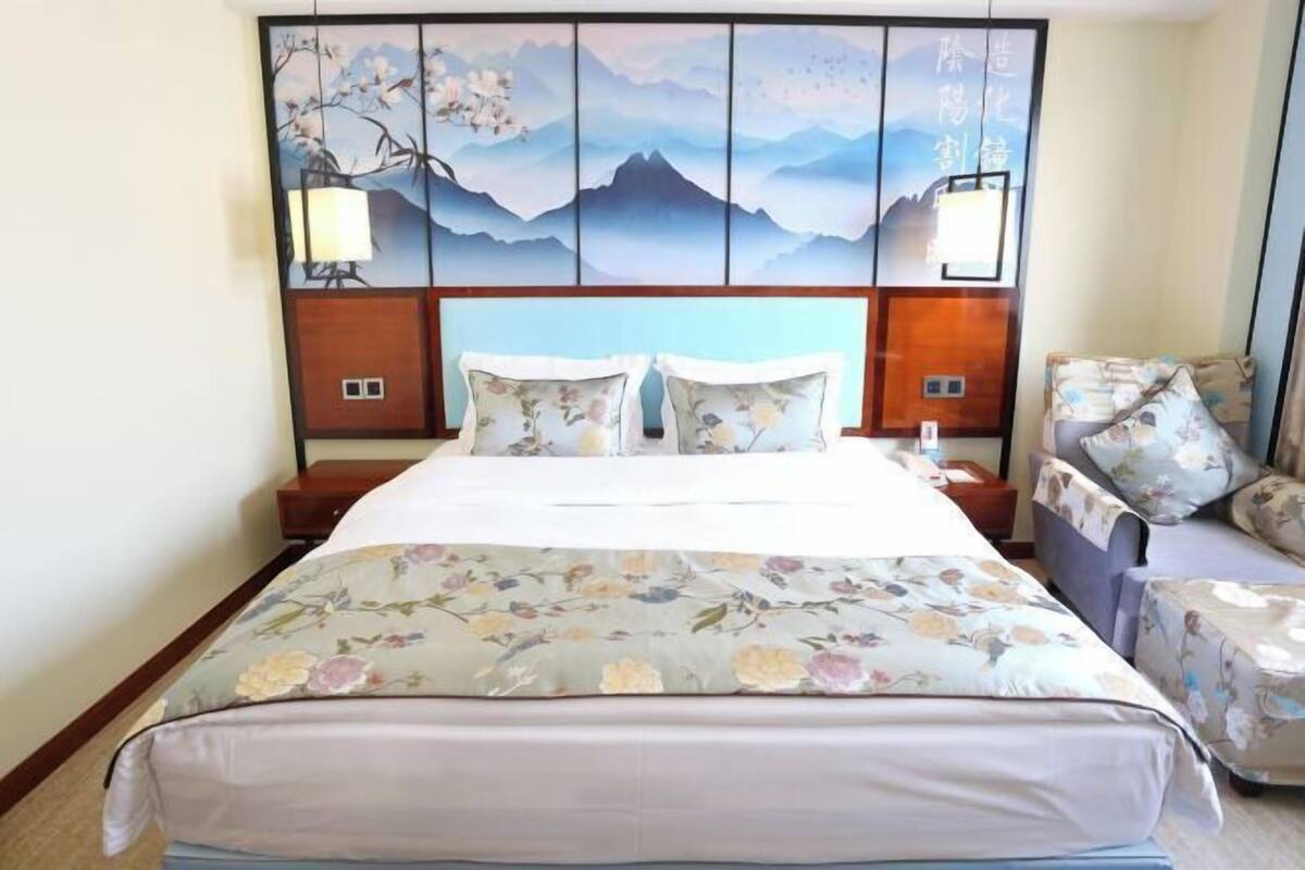 Quarto de hotel com uma cama, roupa de cama florida, cabeceira com arte, mesas de cabeceira e uma cadeira.