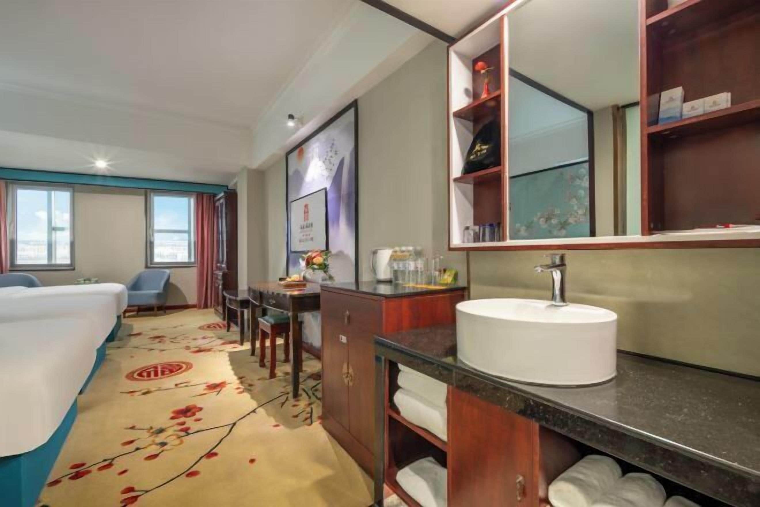A hotel bathroom with a large mirror, a white sink, a cabinet with towels, and a floral carpet.