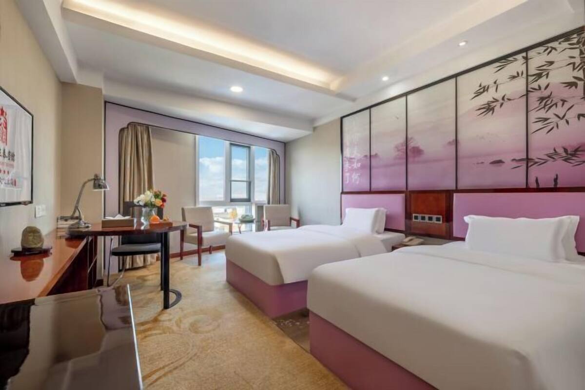 Quarto de hotel com duas camas, uma escrivaninha, uma cadeira, uma janela e um painel decorativo na parede.
