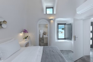 A modern hotel room with a bed, a small table with a plant, a bathroom visible through an archway, and a TV mounted on the wall. - 3 Bedroom Villa with Outdoor Hot Tub & Sea View, Aleanna Traditional Villa (Santorini)