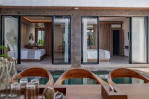 A modern hotel room with a glass door leading to a pool, a separate bedroom, and a dining area with a table and chairs. - Alima Villa Ubud (Sukawati)