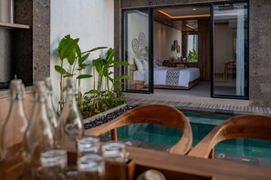 A modern hotel room with a glass door leading to a bedroom, a pool area with wooden chairs, and a dining table with bottles and glasses. - Alima Villa Ubud (Sukawati)