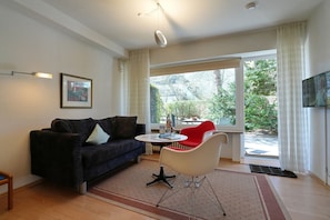 A living room with a dark sofa, a red chair, a small table, and a large window with curtains.