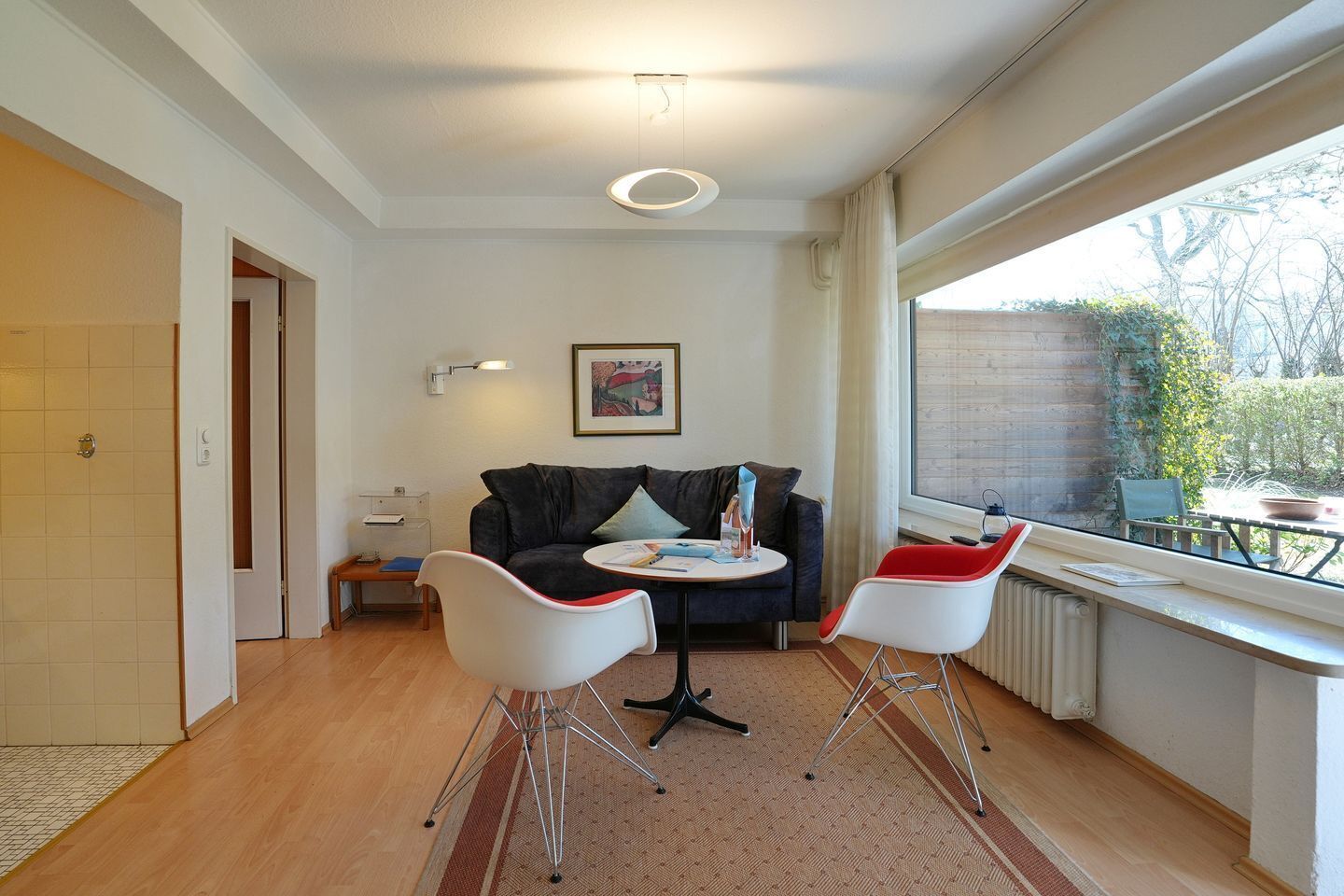 A modern living room with a sofa, two chairs, a small table, a painting, and a window with a view of greenery.