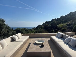 Eine Dachterrasse mit einer beigen Sofagarnitur, einem Tisch in der Mitte mit einer Schale und Blick auf Meer und Hügel.