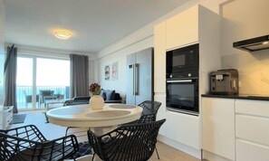 A modern kitchen with a built-in oven, a coffee machine, and a dining area with a round table and chairs. - Elisa - Panoramic View (Saranda)