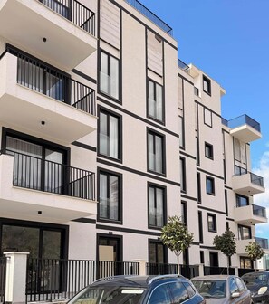 A multi-story residential building with balconies, large windows, and a parking area with cars. - Elisa - Panoramic View (Saranda)